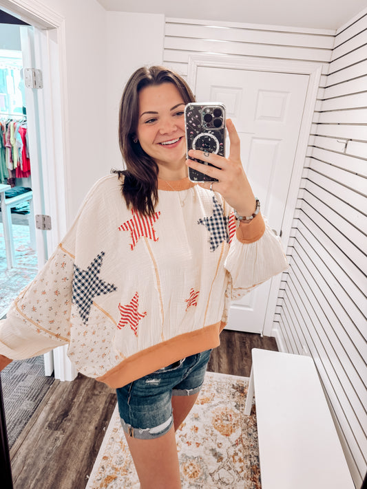 Woman wearing a cream textured pullover with raw seam detailing and patchwork star appliqués in red stripes and blue gingham. Features blush pink ribbed cuffs and hem, floral-print sleeves, and a relaxed, oversized fit. Styled with distressed light-wash denim shorts and a wide-brim cream hat, this cozy top delivers vintage Americana vibes with a soft, boho twist—perfect for patriotic holidays or laid-back summer days.