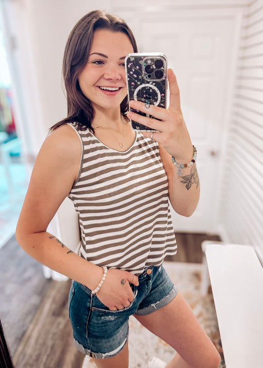 Woman wearing a casual sleeveless tank top with horizontal taupe and white stripes. The top features a scoop neckline and a relaxed, slightly flared silhouette that offers a breezy, comfortable fit. Styled with distressed dark-wash denim shorts, this easygoing look is perfect for everyday wear, beach days, or layering under summer cardigans and jackets.