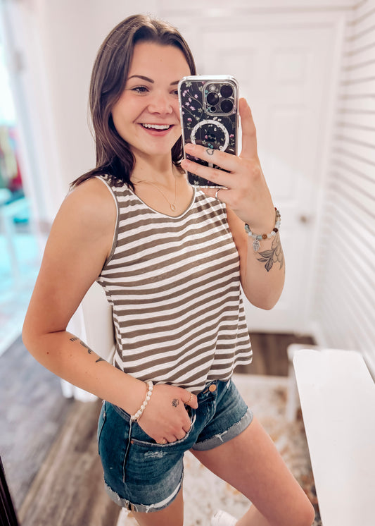 Woman wearing a casual sleeveless tank top with horizontal taupe and white stripes. The top features a scoop neckline and a relaxed, slightly flared silhouette that offers a breezy, comfortable fit. Styled with distressed dark-wash denim shorts, this easygoing look is perfect for everyday wear, beach days, or layering under summer cardigans and jackets.