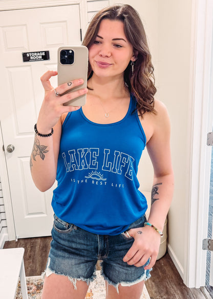 Woman wearing a royal blue racerback tank top with bold white lettering that reads “LAKE LIFE” across the chest and “is the best life” underneath with a small sun graphic. The tank has a relaxed fit and a soft, stretchy fabric, perfect for warm weather and laid-back summer days. She pairs it with dark-wash, frayed denim shorts for a casual, outdoorsy vibe ideal for weekends at the lake, boating, or bonfires.