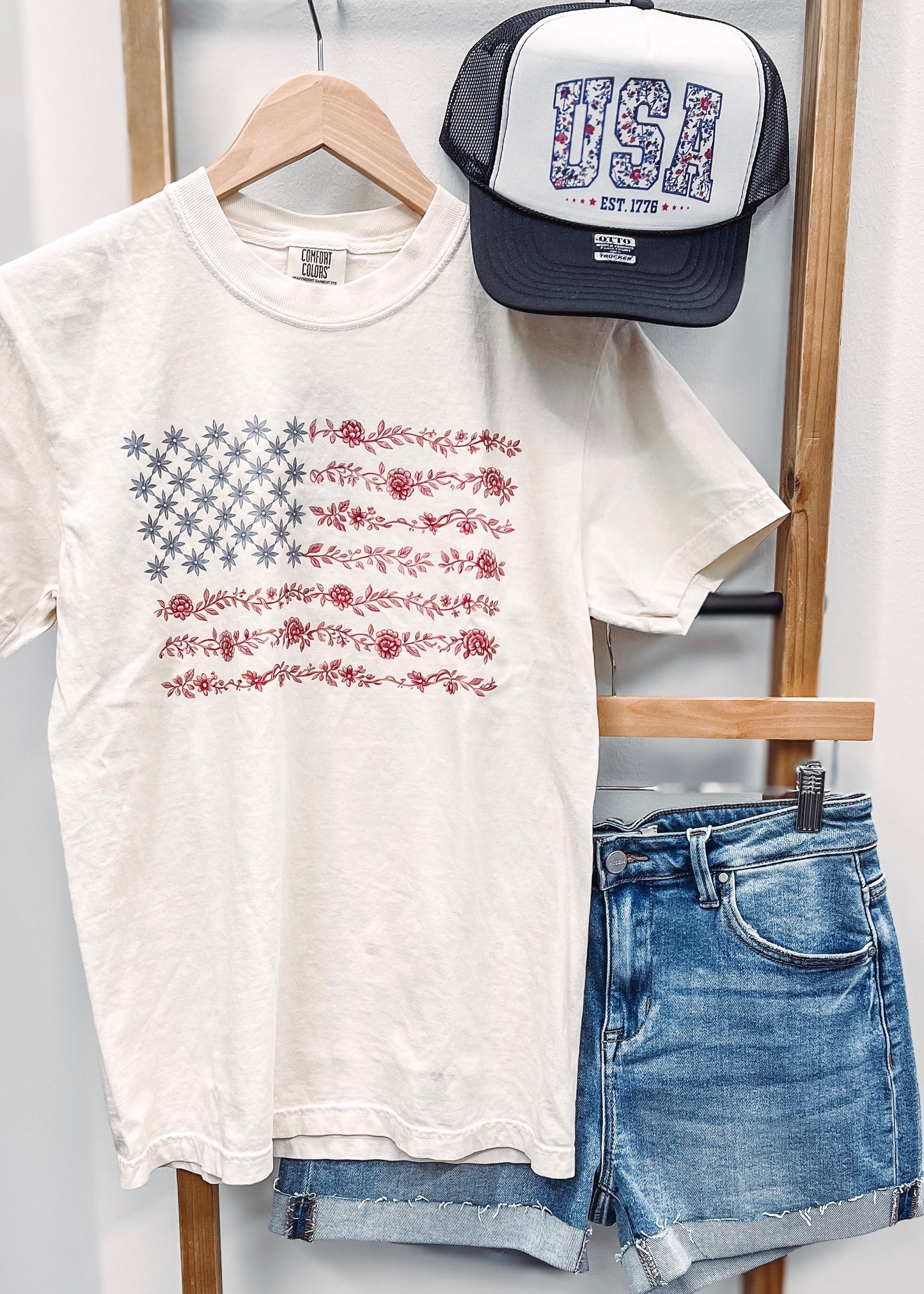 Cream-colored unisex t-shirt featuring a subtle and stylish American flag design made entirely of florals. The star field is composed of blue star-shaped flowers, while the red stripes are formed from intricate red floral garlands. This soft, vintage-inspired tee blends patriotic pride with a feminine, boho twist—perfect for summer celebrations like the Fourth of July, concerts, or casual weekends.