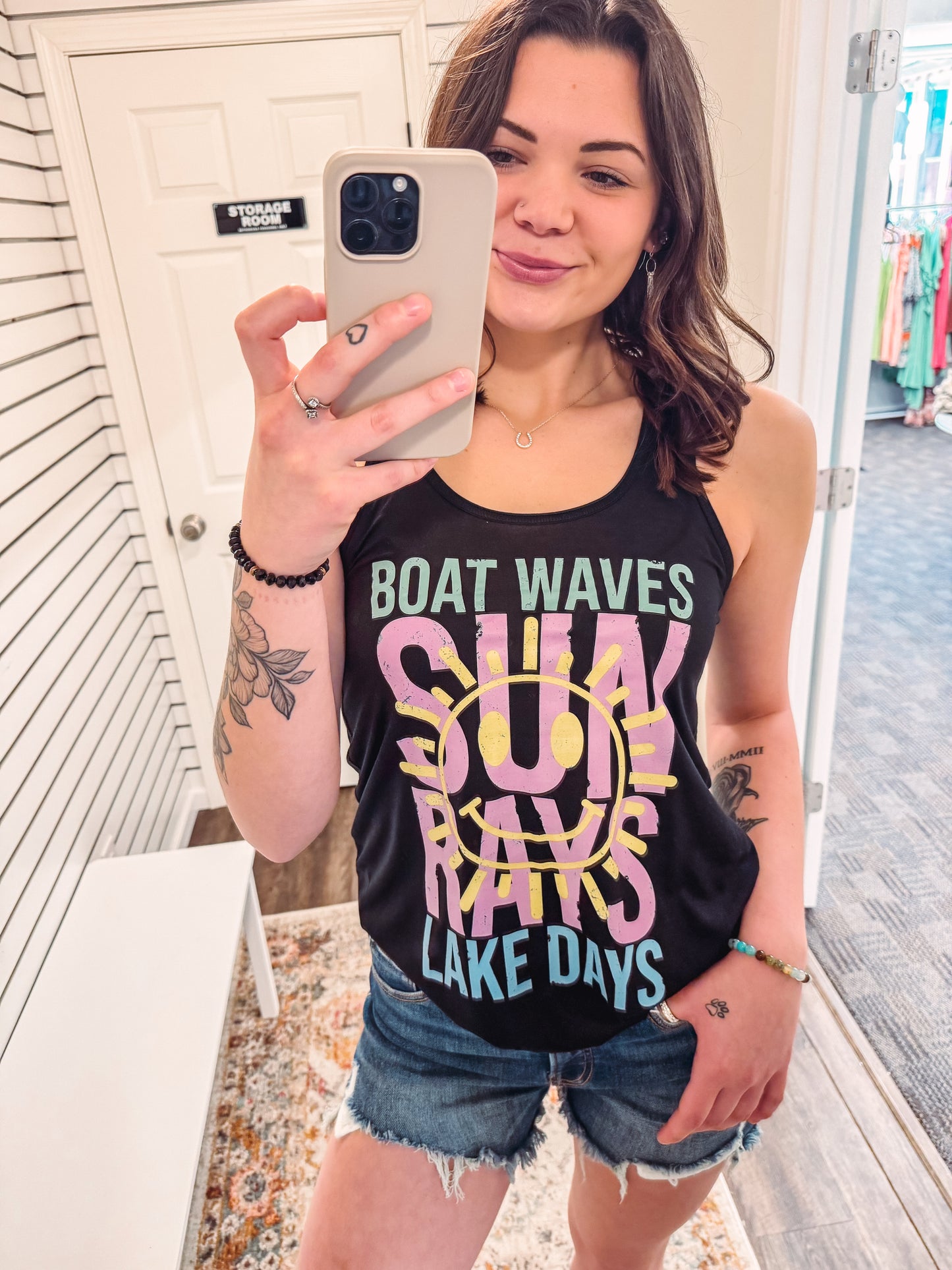 Woman wearing a black racerback tank top with bold, colorful summer-themed text that reads: “BOAT WAVES, SUN RAYS, LAKE DAYS.” The design features a large yellow smiley face sun graphic overlaying the words "SUN RAYS" in pink, with aqua and mint green accents on the other phrases. The tank has a relaxed fit and is paired with frayed denim shorts—perfect for lake weekends, boat rides, or soaking up sunshine in playful, retro style.