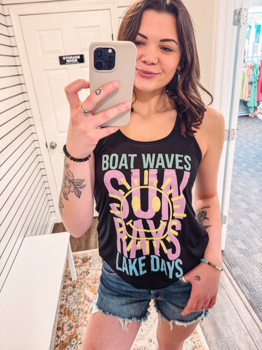 Woman wearing a black racerback tank top with bold, colorful summer-themed text that reads: “BOAT WAVES, SUN RAYS, LAKE DAYS.” The design features a large yellow smiley face sun graphic overlaying the words "SUN RAYS" in pink, with aqua and mint green accents on the other phrases. The tank has a relaxed fit and is paired with frayed denim shorts—perfect for lake weekends, boat rides, or soaking up sunshine in playful, retro style.