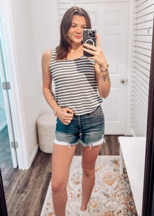 Woman wearing a casual sleeveless tank top with horizontal forest green and white stripes. The top features a scoop neckline and a relaxed, slightly flared silhouette that offers a breezy, comfortable fit. Styled with distressed dark-wash denim shorts, this easygoing look is perfect for everyday wear, beach days, or layering under summer cardigans and jackets.