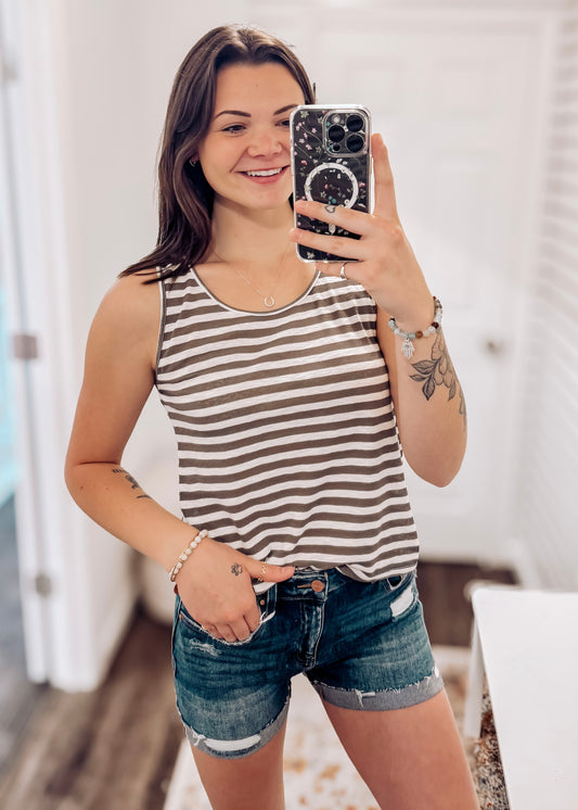 Woman wearing a casual sleeveless tank top with horizontal taupe and white stripes. The top features a scoop neckline and a relaxed, slightly flared silhouette that offers a breezy, comfortable fit. Styled with distressed dark-wash denim shorts, this easygoing look is perfect for everyday wear, beach days, or layering under summer cardigans and jackets.