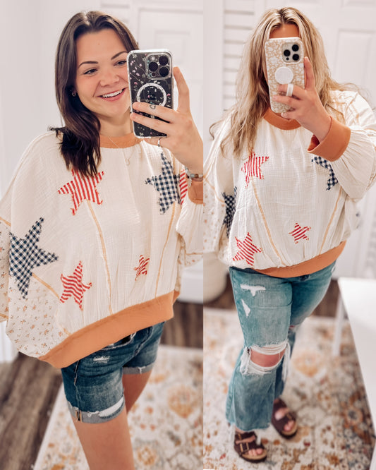 Woman wearing a cream textured pullover with raw seam detailing and patchwork star appliqués in red stripes and blue gingham. Features blush pink ribbed cuffs and hem, floral-print sleeves, and a relaxed, oversized fit. Styled with distressed light-wash denim shorts and a wide-brim cream hat, this cozy top delivers vintage Americana vibes with a soft, boho twist—perfect for patriotic holidays or laid-back summer days.