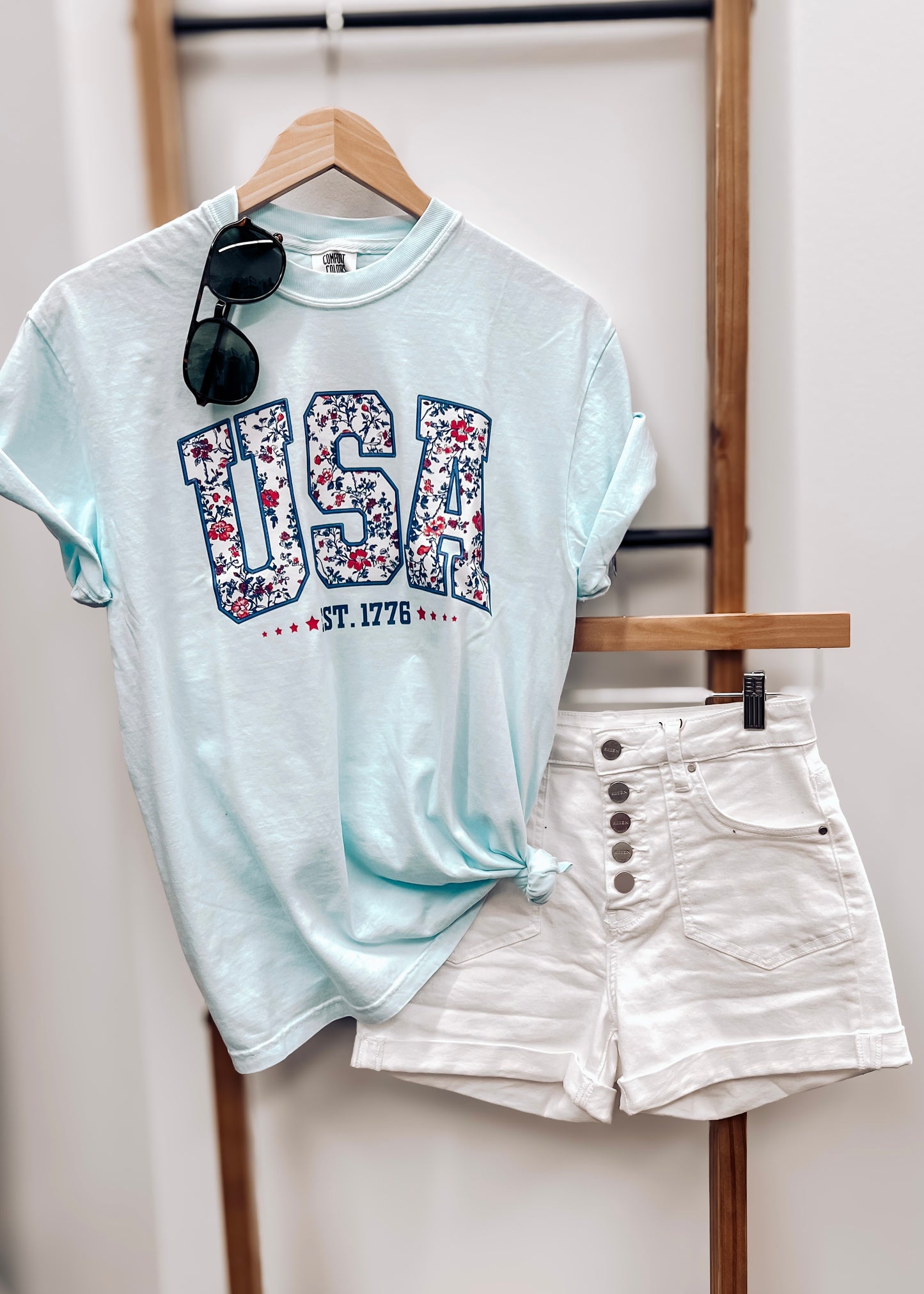 Light blue unisex t-shirt featuring bold "USA" block letters filled with a red, white, and blue floral pattern for a faux-embroidered look. Below, "EST. 1776" is printed in navy with red stars, celebrating American heritage. Classic crew neck and short sleeves offer a comfortable fit, perfect for patriotic holidays like the Fourth of July or Memorial Day.