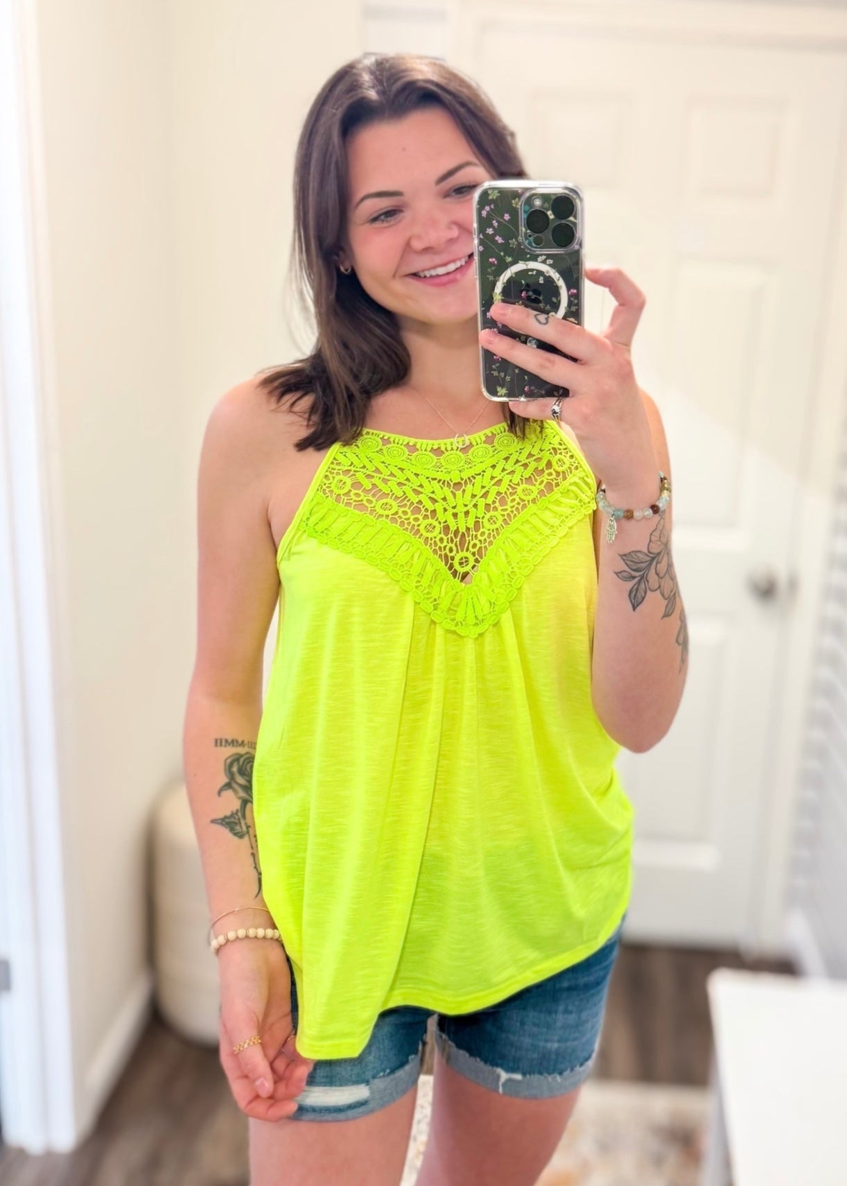 Woman taking a mirror selfie wearing a neon yellow sleeveless halter tank top with intricate crochet lace detailing along the neckline and chest. The flowy, lightweight fabric offers a relaxed fit, perfect for summer. She pairs it with medium-wash denim shorts. The vibrant color and breezy design make it an eye-catching, warm-weather staple.