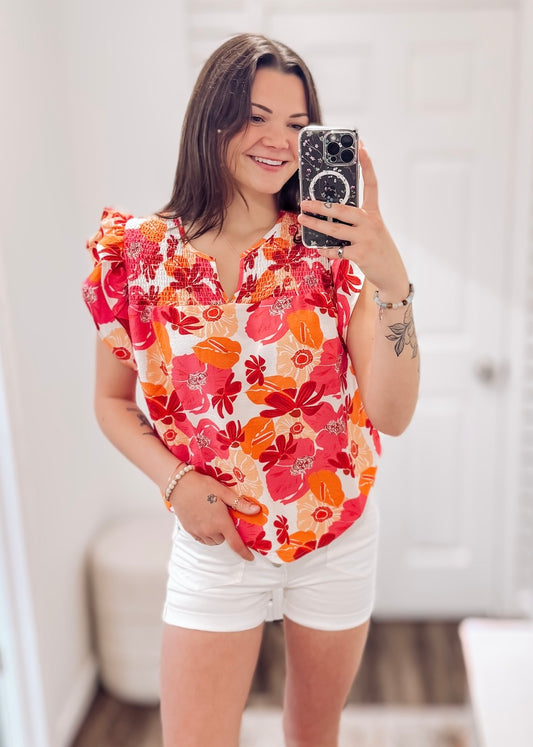 Woman wearing a vibrant floral short-sleeve top with a textured, crinkle fabric. The blouse features shades of pink, red, orange, and white in a bold floral print, with ruffled sleeves and a smocked detail across the chest. A split V-neckline adds a flattering touch, and the relaxed fit pairs effortlessly with her high-rise white distressed denim shorts. A bright, cheerful piece perfect for summer days, vacations, or casual outings.