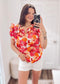 Woman wearing a vibrant floral short-sleeve top with a textured, crinkle fabric. The blouse features shades of pink, red, orange, and white in a bold floral print, with ruffled sleeves and a smocked detail across the chest. A split V-neckline adds a flattering touch, and the relaxed fit pairs effortlessly with her high-rise white distressed denim shorts. A bright, cheerful piece perfect for summer days, vacations, or casual outings.