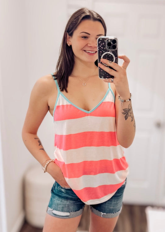 Woman wearing a sleeveless pink and white striped tank top with a relaxed, flowy fit. The top features bold horizontal stripes and a deep V-neckline trimmed in aqua blue, adding a fun pop of contrast. Thin spaghetti straps give it a breezy, beachy vibe, perfect for warm weather. Paired with distressed light-wash denim shorts, this playful tank is ideal for casual summer days, festivals, or vacation looks.