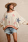 Woman wearing a cream textured pullover with raw seam detailing and patchwork star appliqués in red stripes and blue gingham. Features blush pink ribbed cuffs and hem, floral-print sleeves, and a relaxed, oversized fit. Styled with distressed light-wash denim shorts and a wide-brim cream hat, this cozy top delivers vintage Americana vibes with a soft, boho twist—perfect for patriotic holidays or laid-back summer days.