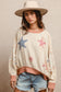Woman wearing a cream textured pullover with raw seam detailing and patchwork star appliqués in red stripes and blue gingham. Features blush pink ribbed cuffs and hem, floral-print sleeves, and a relaxed, oversized fit. Styled with distressed light-wash denim shorts and a wide-brim cream hat, this cozy top delivers vintage Americana vibes with a soft, boho twist—perfect for patriotic holidays or laid-back summer days.