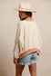 Woman wearing a cream textured pullover with raw seam detailing and patchwork star appliqués in red stripes and blue gingham. Features blush pink ribbed cuffs and hem, floral-print sleeves, and a relaxed, oversized fit. Styled with distressed light-wash denim shorts and a wide-brim cream hat, this cozy top delivers vintage Americana vibes with a soft, boho twist—perfect for patriotic holidays or laid-back summer days.