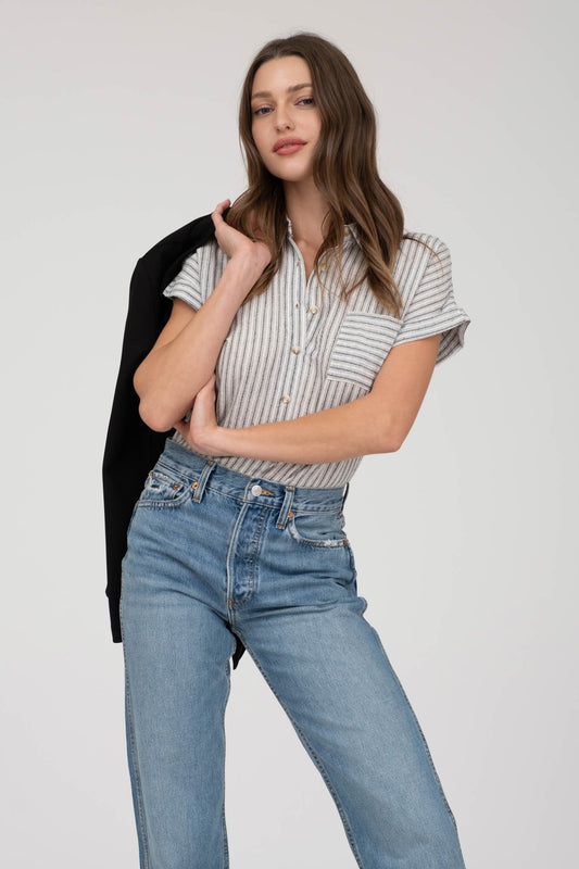 model wearing a short-sleeve button-up top with vertical black and white pinstripes. Features a collared neckline, front chest pocket with horizontal stripes for contrast, and a relaxed, slightly oversized fit. The shirt is styled with a black denim mini skirt, creating an effortlessly chic and casual look perfect for everyday wear