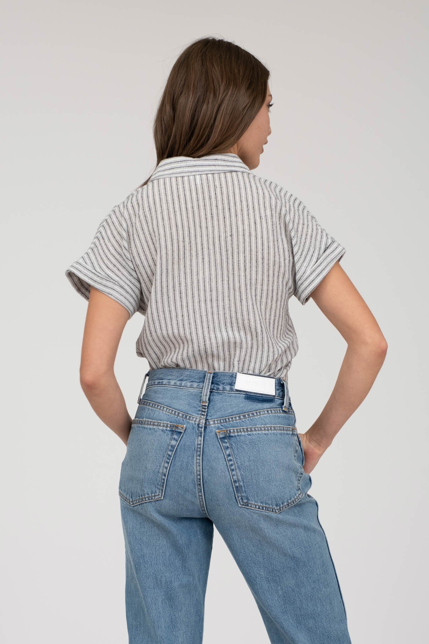 model wearing a short-sleeve button-up top with vertical black and white pinstripes. Features a collared neckline, front chest pocket with horizontal stripes for contrast, and a relaxed, slightly oversized fit. The shirt is styled with a black denim mini skirt, creating an effortlessly chic and casual look perfect for everyday wear