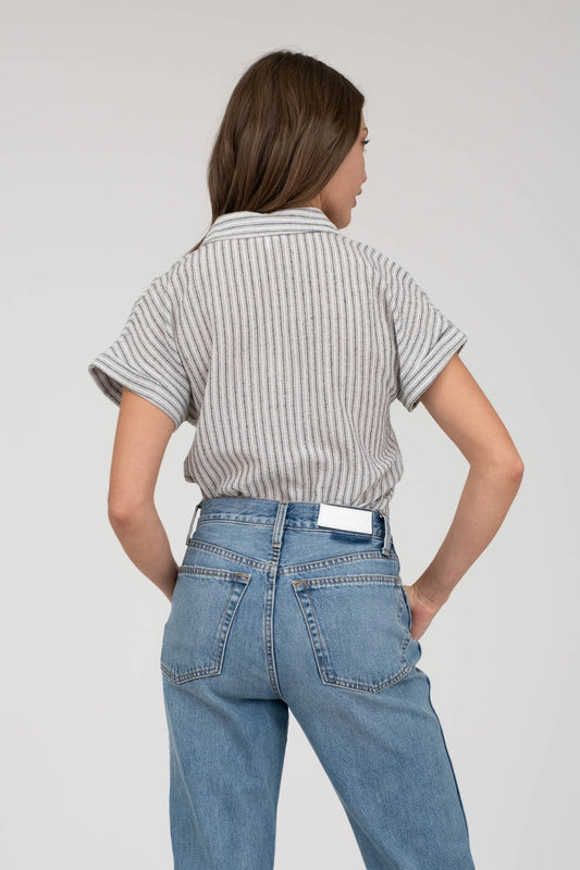 model wearing a short-sleeve button-up top with vertical black and white pinstripes. Features a collared neckline, front chest pocket with horizontal stripes for contrast, and a relaxed, slightly oversized fit. The shirt is styled with a black denim mini skirt, creating an effortlessly chic and casual look perfect for everyday wear