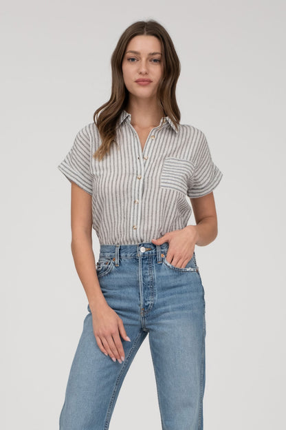 model wearing a short-sleeve button-up top with vertical black and white pinstripes. Features a collared neckline, front chest pocket with horizontal stripes for contrast, and a relaxed, slightly oversized fit. The shirt is styled with a black denim mini skirt, creating an effortlessly chic and casual look perfect for everyday wear