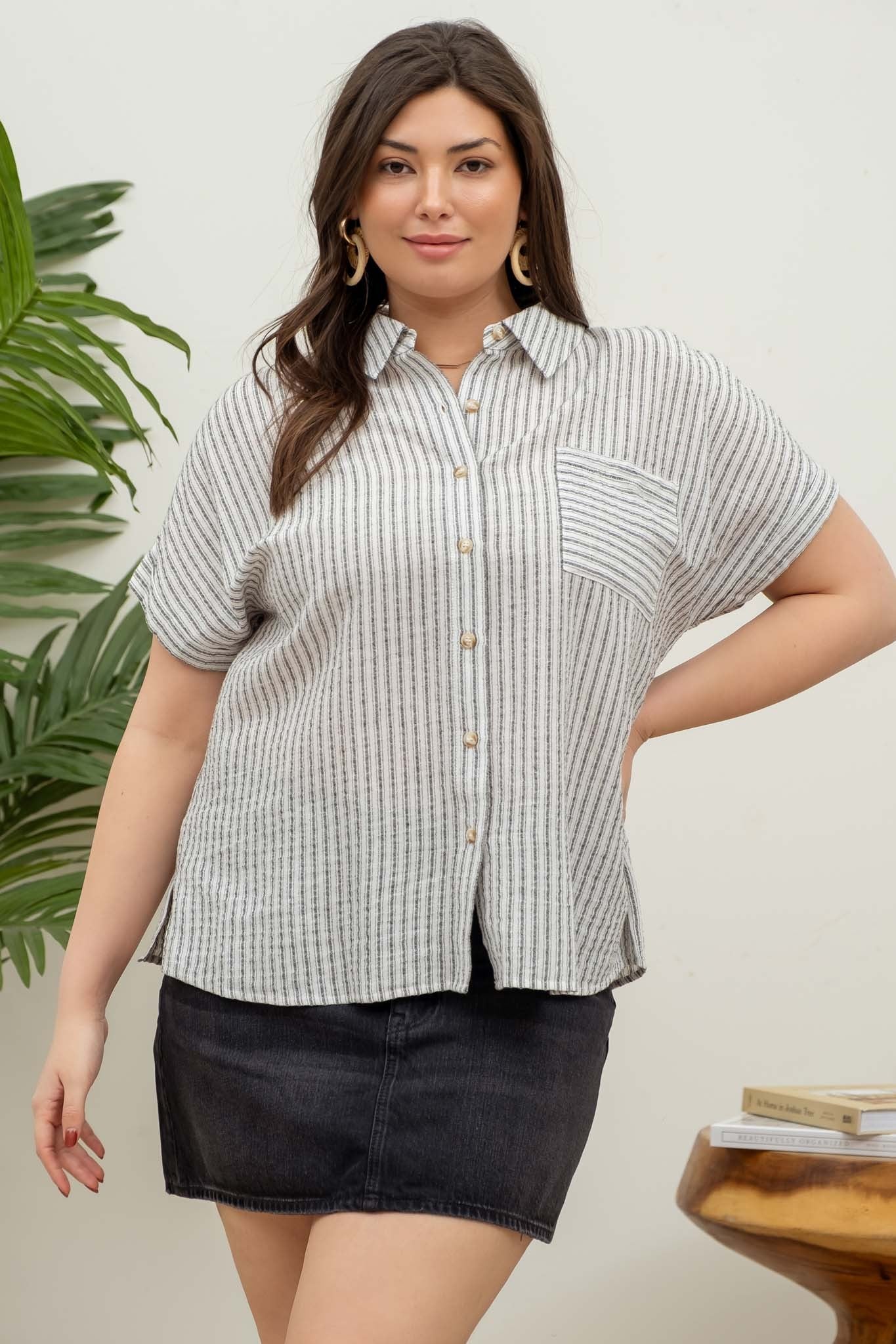 model wearing a short-sleeve button-up top with vertical black and white pinstripes. Features a collared neckline, front chest pocket with horizontal stripes for contrast, and a relaxed, slightly oversized fit. The shirt is styled with a black denim mini skirt, creating an effortlessly chic and casual look perfect for everyday wear