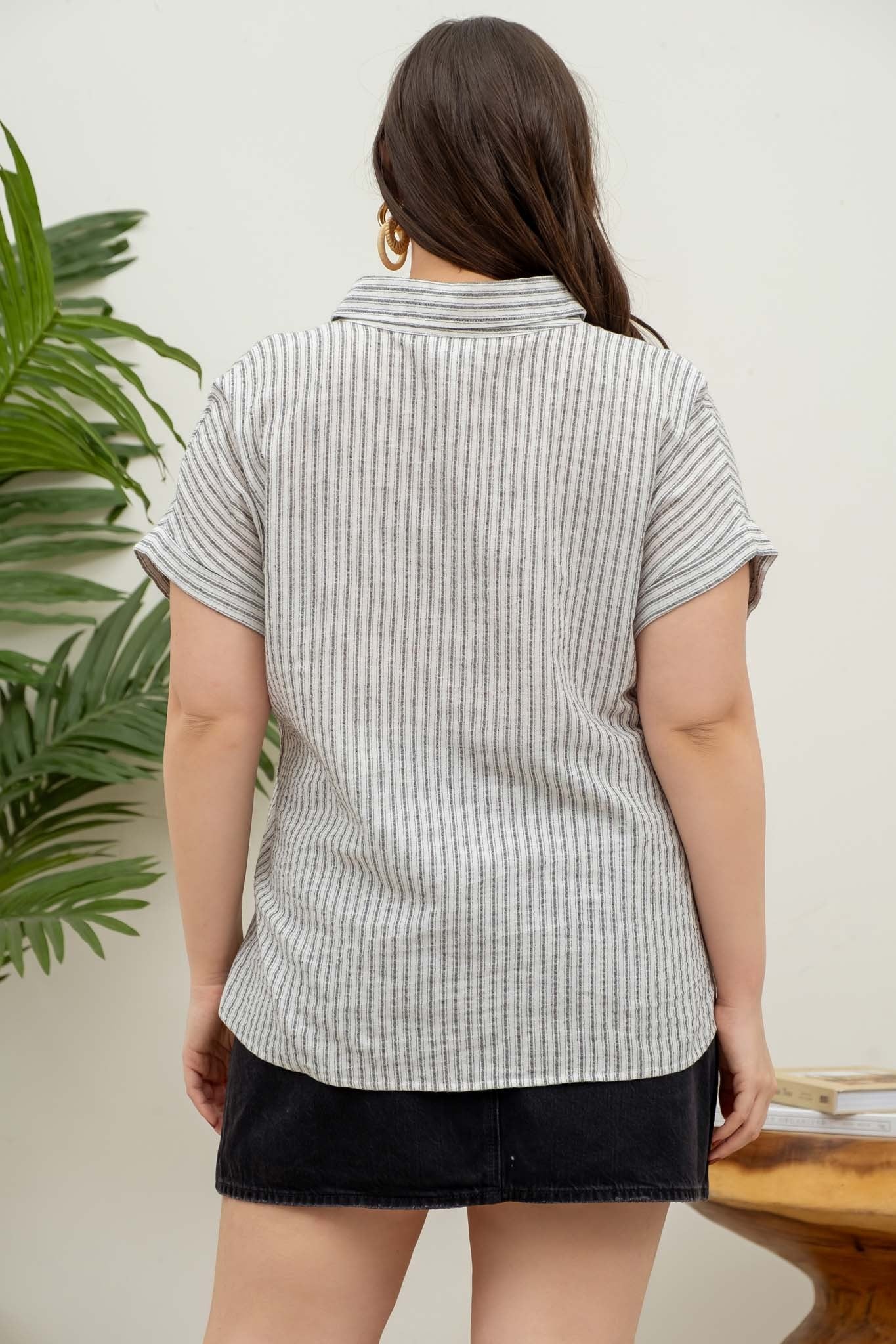 model wearing a short-sleeve button-up top with vertical black and white pinstripes. Features a collared neckline, front chest pocket with horizontal stripes for contrast, and a relaxed, slightly oversized fit. The shirt is styled with a black denim mini skirt, creating an effortlessly chic and casual look perfect for everyday wear