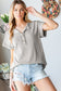 Woman wearing a casual, light taupe short-sleeve henley-style top with a soft ribbed texture and two-button placket neckline. The top features a relaxed, slightly oversized fit with rolled sleeves and a subtle high-low hem. She pairs it with light wash distressed denim shorts that have heavy fraying and large rips along the front. Her long, wavy blonde hair is styled down, and she poses indoors near spring floral decor and a pastel flower wreath on the wall. The setting and styling give off an effortless, c