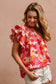 Woman wearing a vibrant floral short-sleeve top with a textured, crinkle fabric. The blouse features shades of pink, red, orange, and white in a bold floral print, with ruffled sleeves and a smocked detail across the chest. A split V-neckline adds a flattering touch, and the relaxed fit pairs effortlessly with her high-rise white distressed denim shorts. A bright, cheerful piece perfect for summer days, vacations, or casual outings.