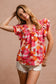 Woman wearing a vibrant floral short-sleeve top with a textured, crinkle fabric. The blouse features shades of pink, red, orange, and white in a bold floral print, with ruffled sleeves and a smocked detail across the chest. A split V-neckline adds a flattering touch, and the relaxed fit pairs effortlessly with her high-rise white distressed denim shorts. A bright, cheerful piece perfect for summer days, vacations, or casual outings.