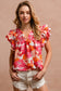 Woman wearing a vibrant floral short-sleeve top with a textured, crinkle fabric. The blouse features shades of pink, red, orange, and white in a bold floral print, with ruffled sleeves and a smocked detail across the chest. A split V-neckline adds a flattering touch, and the relaxed fit pairs effortlessly with her high-rise white distressed denim shorts. A bright, cheerful piece perfect for summer days, vacations, or casual outings.
