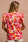 Woman wearing a vibrant floral short-sleeve top with a textured, crinkle fabric. The blouse features shades of pink, red, orange, and white in a bold floral print, with ruffled sleeves and a smocked detail across the chest. A split V-neckline adds a flattering touch, and the relaxed fit pairs effortlessly with her high-rise white distressed denim shorts. A bright, cheerful piece perfect for summer days, vacations, or casual outings.