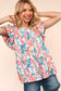 Woman wearing a vibrant, multicolored floral print babydoll top with a round neckline and flutter sleeves. The top features a loose, flowy silhouette with a gathered waist seam that adds volume and movement. The pattern includes shades of coral, teal, pink, orange, and blue against a white background, creating a playful, summery look. She pairs it with medium-wash denim shorts. Her long blonde hair is styled in soft waves, and she stands against a light peach backdrop that complements the warm tones of the 