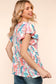 Woman wearing a vibrant, multicolored floral print babydoll top with a round neckline and flutter sleeves. The top features a loose, flowy silhouette with a gathered waist seam that adds volume and movement. The pattern includes shades of coral, teal, pink, orange, and blue against a white background, creating a playful, summery look. She pairs it with medium-wash denim shorts. Her long blonde hair is styled in soft waves, and she stands against a light peach backdrop that complements the warm tones of the 