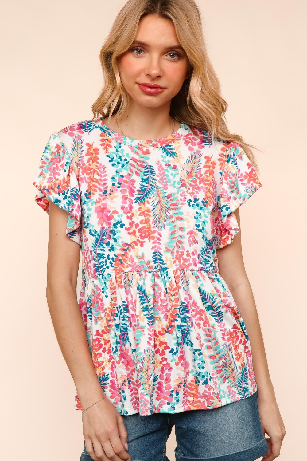 Woman wearing a vibrant, multicolored floral print babydoll top with a round neckline and flutter sleeves. The top features a loose, flowy silhouette with a gathered waist seam that adds volume and movement. The pattern includes shades of coral, teal, pink, orange, and blue against a white background, creating a playful, summery look. She pairs it with medium-wash denim shorts. Her long blonde hair is styled in soft waves, and she stands against a light peach backdrop that complements the warm tones of the 