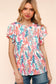 Woman wearing a vibrant, multicolored floral print babydoll top with a round neckline and flutter sleeves. The top features a loose, flowy silhouette with a gathered waist seam that adds volume and movement. The pattern includes shades of coral, teal, pink, orange, and blue against a white background, creating a playful, summery look. She pairs it with medium-wash denim shorts. Her long blonde hair is styled in soft waves, and she stands against a light peach backdrop that complements the warm tones of the 