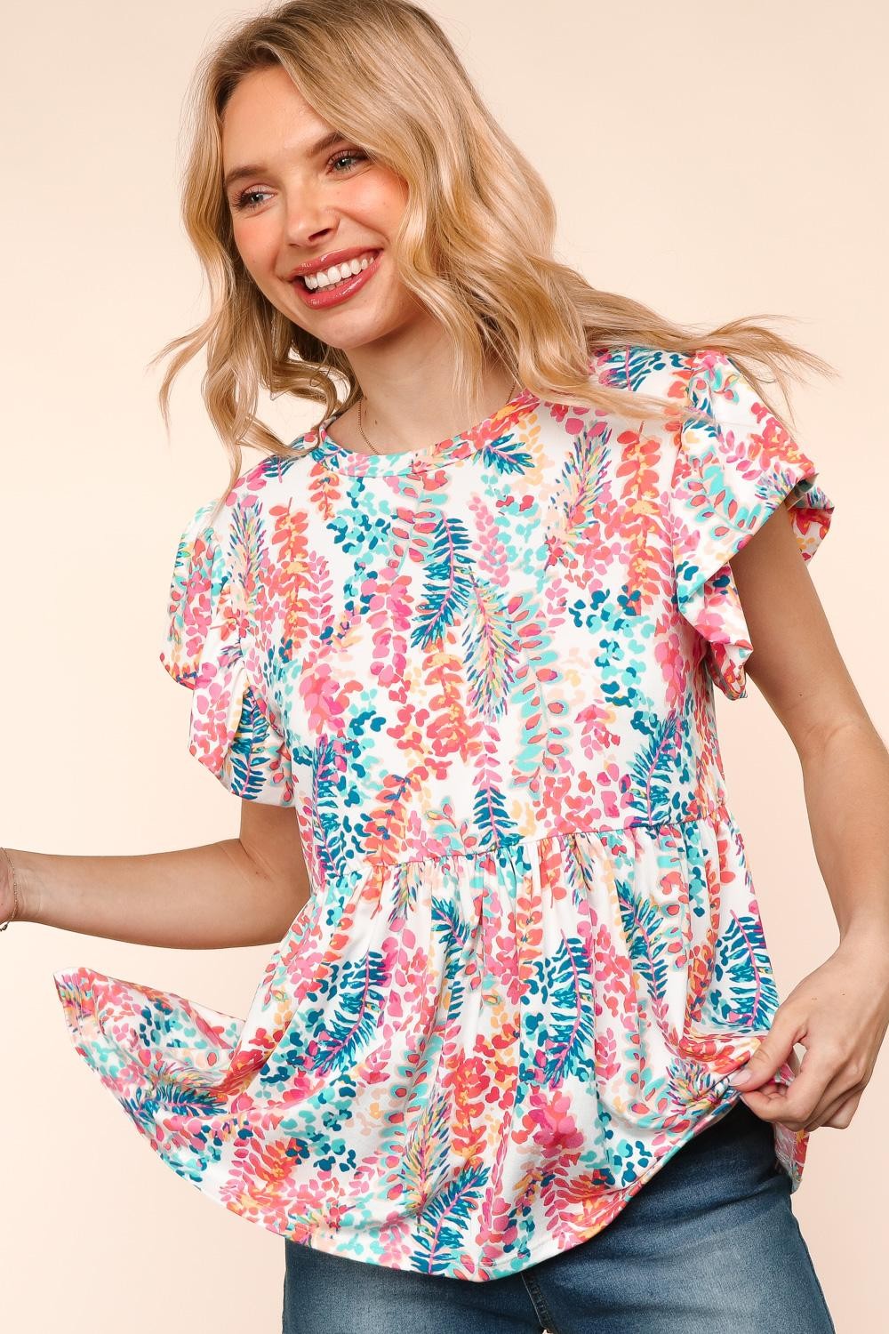 Woman wearing a vibrant, multicolored floral print babydoll top with a round neckline and flutter sleeves. The top features a loose, flowy silhouette with a gathered waist seam that adds volume and movement. The pattern includes shades of coral, teal, pink, orange, and blue against a white background, creating a playful, summery look. She pairs it with medium-wash denim shorts. Her long blonde hair is styled in soft waves, and she stands against a light peach backdrop that complements the warm tones of the 