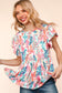 Woman wearing a vibrant, multicolored floral print babydoll top with a round neckline and flutter sleeves. The top features a loose, flowy silhouette with a gathered waist seam that adds volume and movement. The pattern includes shades of coral, teal, pink, orange, and blue against a white background, creating a playful, summery look. She pairs it with medium-wash denim shorts. Her long blonde hair is styled in soft waves, and she stands against a light peach backdrop that complements the warm tones of the 